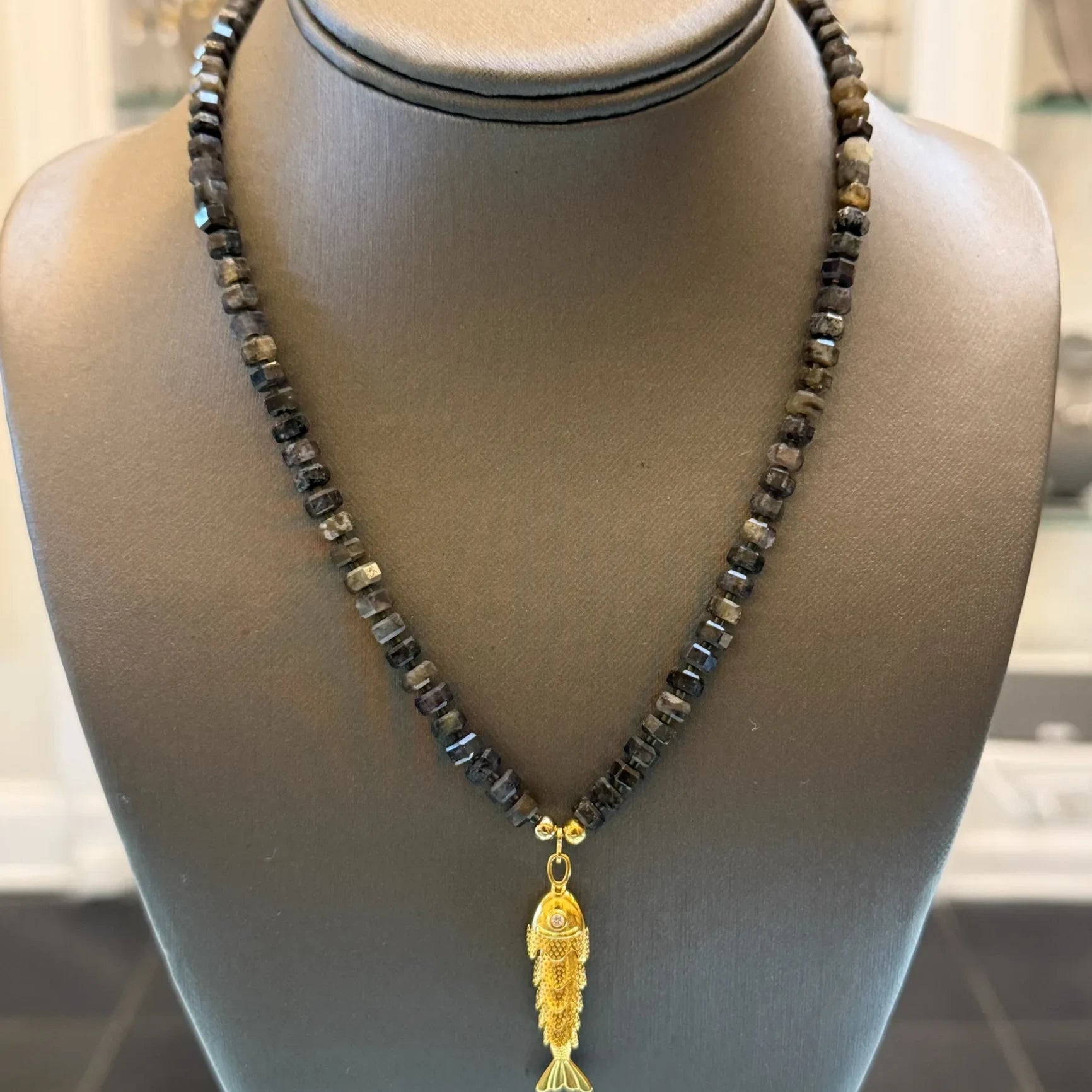 CHAKARR beaded necklace featuring a gold-tone fish pendant with a sparkling accent on faceted dark gemstone beads.