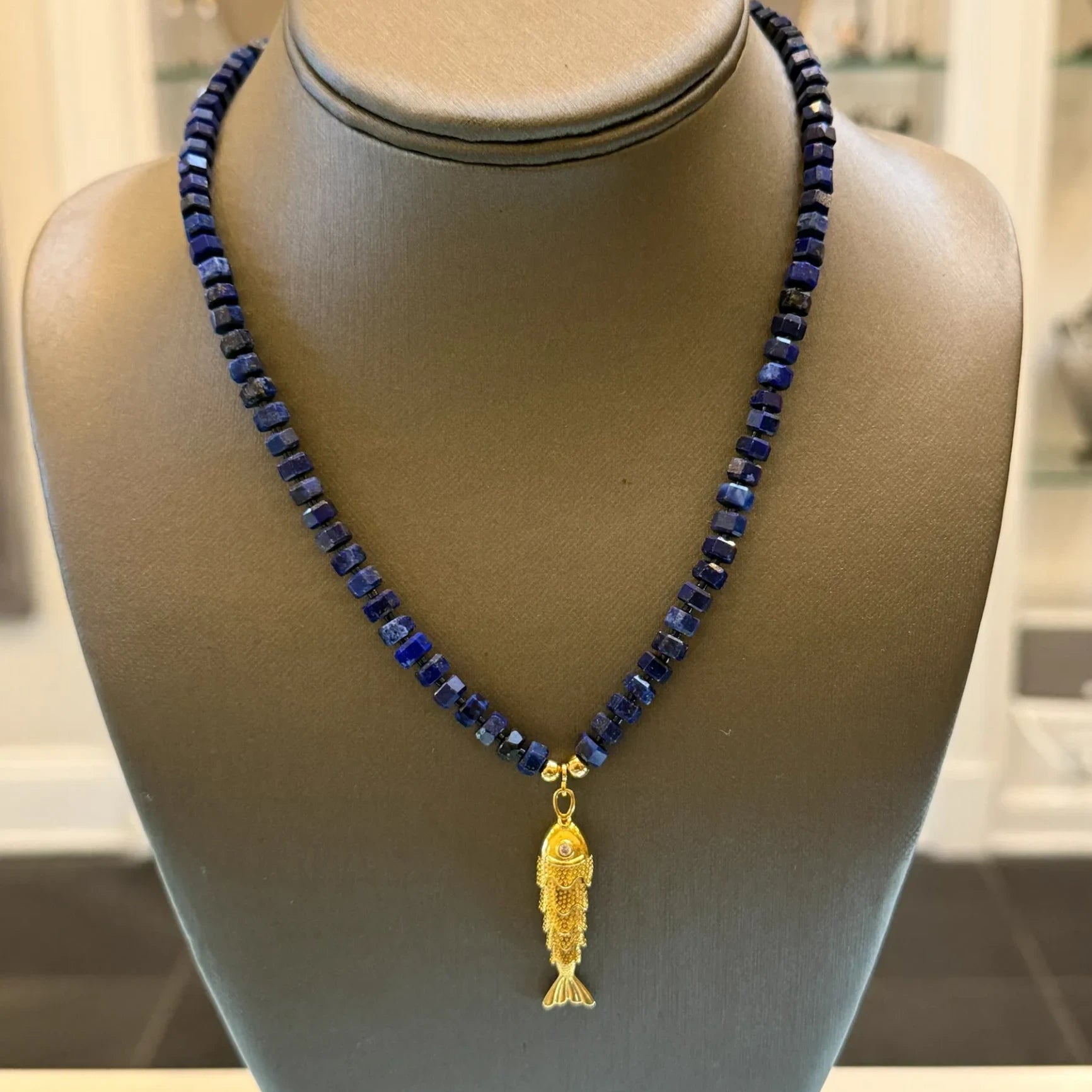 CHAKARR beaded necklace featuring deep blue faceted lapis beads and a gold-tone fish pendant on a display bust.