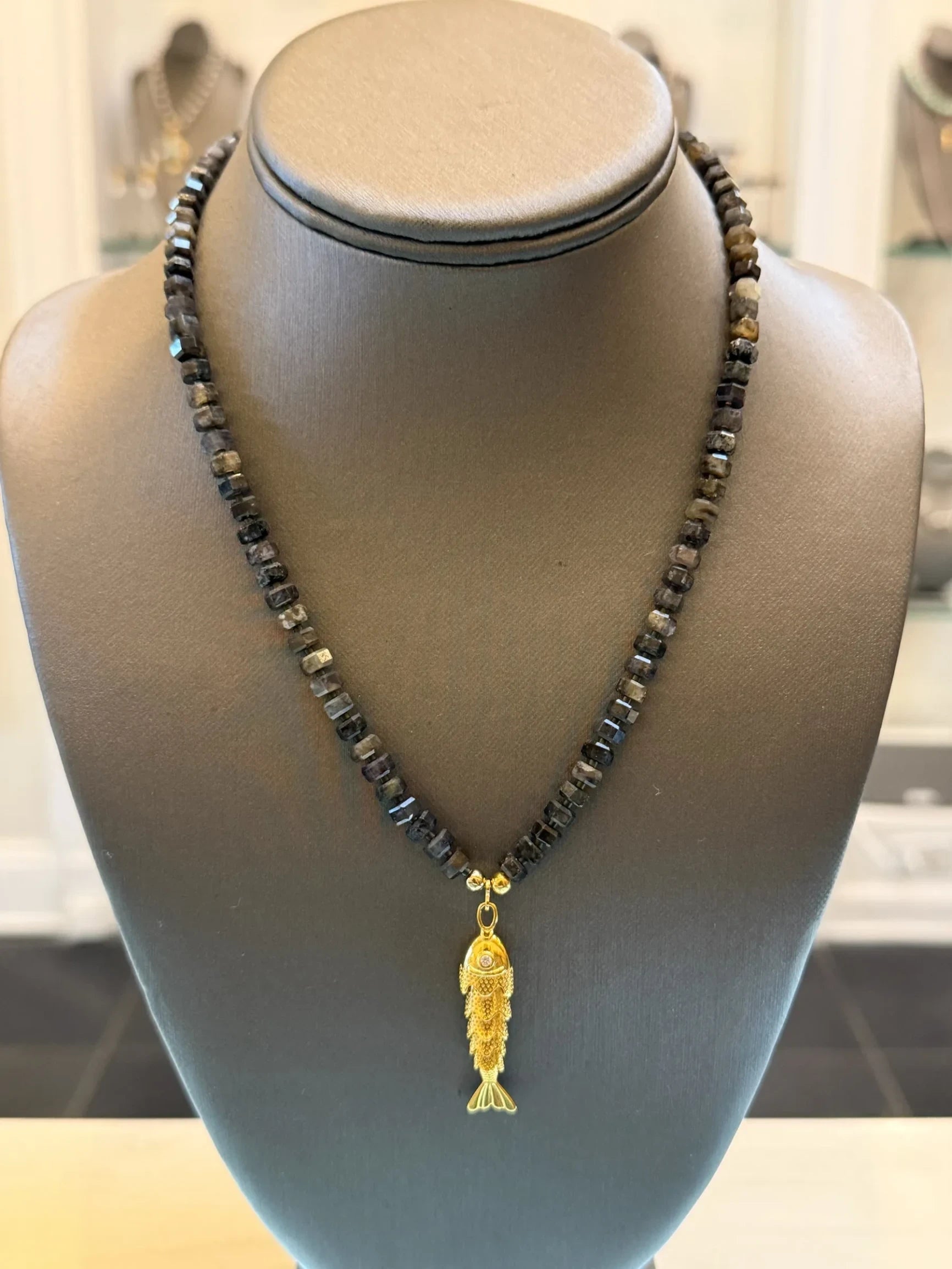 CHAKARR beaded necklace featuring a gold-tone fish pendant with a sparkling accent on faceted dark gemstone beads.