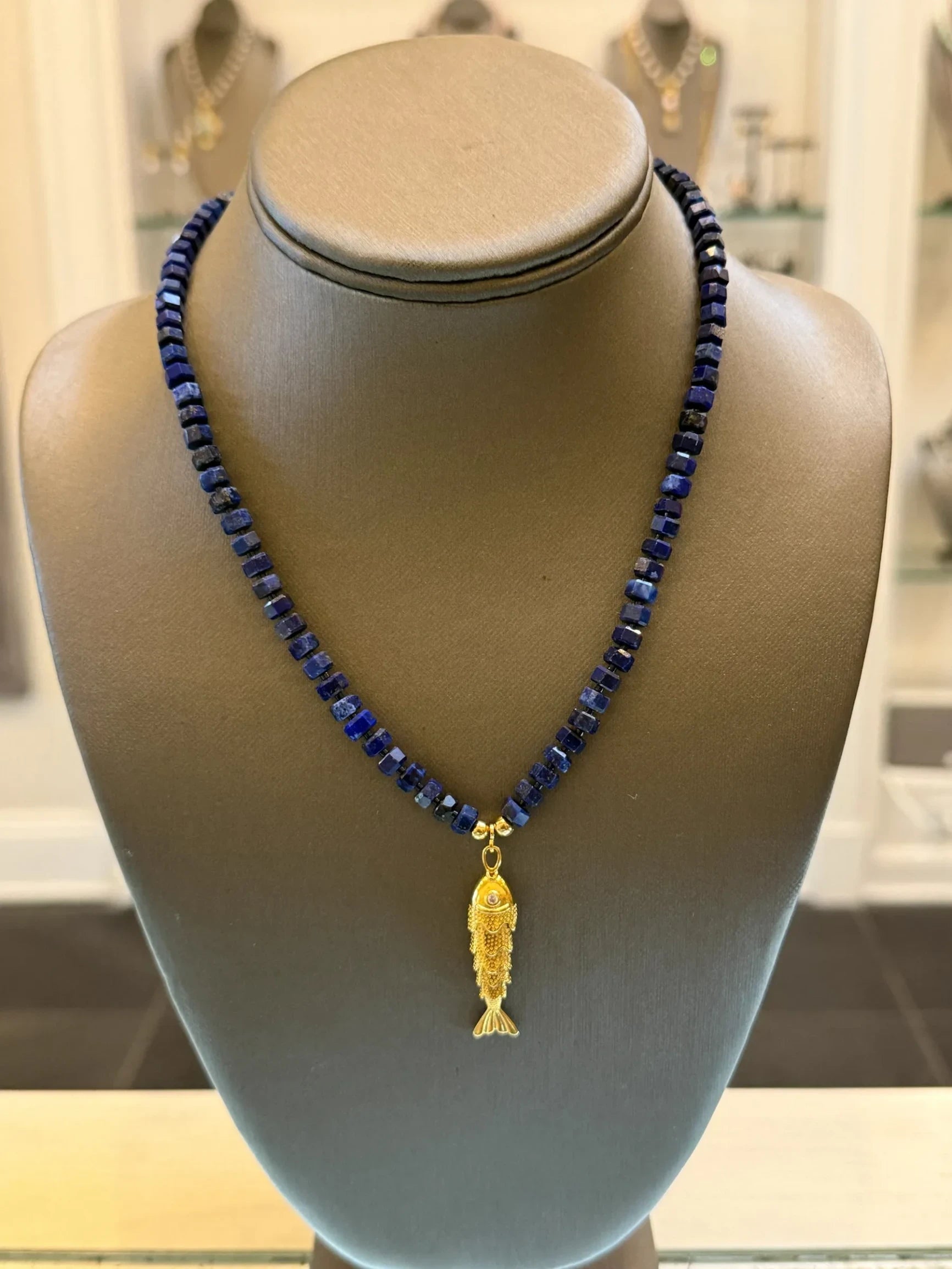 CHAKARR beaded necklace featuring deep blue faceted lapis beads and a gold-tone fish pendant on a display bust.